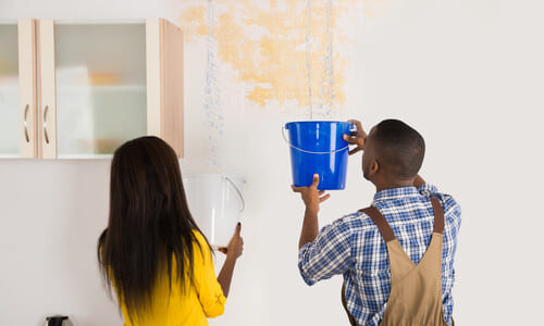 A man and a woman holding buckets up to catch water from a damaged ceiling.