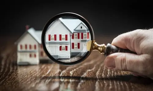 A magnifying glass held in front of a model house placed on a reflective hardwood surface.