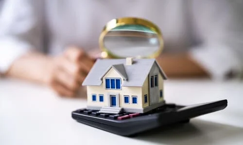 A public adjuster holding a magnifying lens up while looking at a model house on top of a calculator.